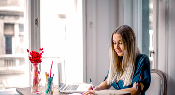 Home-Office: Frau sitzt am Schreibtisch und arbeitet am PC mit einem Buch