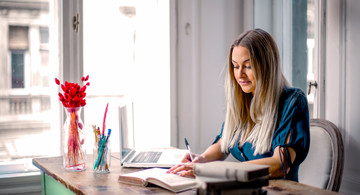 Home-Office: Frau sitzt am Schreibtisch und arbeitet am PC mit einem Buch